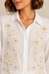Camisa blanca de manga larga confeccionada en lino y viscosa, con cuello camisero, cierre frontal con botones y delicados bordados florales con apliques de perlas y pedrería en el frente.