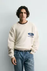 Sudadera de tejido con algodón, color beige claro, de corte relajado (relaxed fit) y cuello redondo. Presenta un parche bordado en el pecho con el logo de Ford y el texto "Genuine Parts" en azul marino.