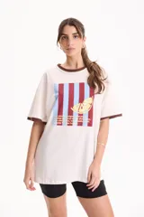 Remera de algodón blanca de manga corta, con corte suelto (loose fit) y rib en contraste marrón en el cuello y los puños. Presenta un estampado frontal rectangular con rayas verticales en tonos burdeos y celestes, superpuesto con el dibujo de una rodaja de limón amarilla y texto impreso.