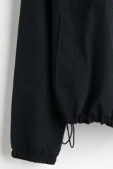 Campera rompevientos de nylon color negro, repelente al agua, con capucha ajustable con cordón, cierre frontal y bolsillos laterales. Cuenta con elásticos en los puños y forro interno.