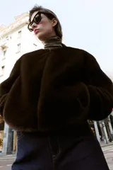 Chaqueta bomber de cuello redondo y manga larga, confeccionada en tejido de pelo sintético color marrón oscuro. Presenta cierre frontal con cremallera, bolsillos laterales y acabados elásticos en el ruedo y puños.