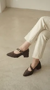 Zapato tipo Mary Jane de gamuza color marrón, con taco cuadrado medio y tira ajustable con hebilla metálica y ojalillos decorativos.