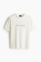Remera blanca de punto de algodón, de corte holgado, con cuello redondo y hombros caídos. Presenta un estampado frontal en color verde con el texto "SEQUOIA PARK" y pequeños iconos alusivos.