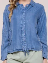 Camperita de denim liviano color azul, con cuello camisero, botones frontales ocultos, mangas largas de hombros caídos, bolsillos laterales y cordón ajustable en la cintura.