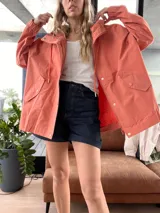 Campera color ladrillo con capucha, cierre frontal con cremallera y broches a presión, bolsillos laterales con solapa y puños elásticos.