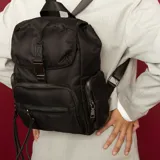 Mochila negra de nylon con tapa y broche metálico. Tiene apertura superior con broche, bolsillos internos, bolsillo con cierre metálico al frente y laterales. Tiene asas rígidas de mano y correas regulables para colgar en la espalda.