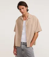Camisa de algodón texturizado color beige, con cuello clásico, mangas cortas y cierre frontal de botones. Presenta un diseño holgado tipo boxy de corte moderno.