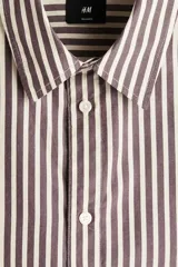 Camisa de algodón suave con cuello inglés, tapeta clásica y canesú en la espalda. Modelo de manga larga con puños abotonados y bajo levemente redondeado. Presenta diseño a rayas en tonos marrón y blanco.