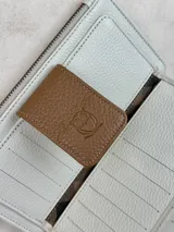 Billetera mediana de cuero color blanco y marrón con cierre de cremallera, compartimentos internos y correa de muñeca.