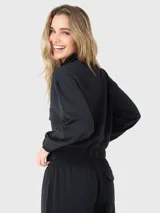 Buzo negro de tela suave y ligera con cuello alto, cierre frontal con botones a presión, bolsillos en la parte frontal y pretina elástica en la cintura.