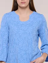 Blusa celeste de tejido texturado con relieve floral, escote redondo con muesca central, mangas tres cuartos estilo campana y ruedo curvo.
