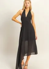Vestido largo negro con escote halter y espalda descubierta. Presenta un diseño asimétrico con una capa superpuesta de tela semitransparente que cae lateralmente, creando un efecto de movimiento y elegancia.