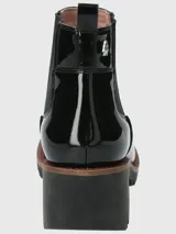 Bota tipo Chelsea de caña corta en charol negro, con paneles laterales elásticos, suela track robusta y taco cuadrado. Presenta un detalle de costura en color rojo en la base de la suela.