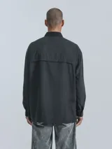 Camisa de manga larga estilo técnico, color negro, con corte oversize. Presenta múltiples bolsillos frontales con solapa y cierre, y un bolsillo adicional en la parte inferior de la espalda. Confeccionada en microfibra suave.