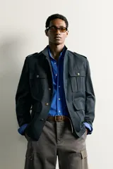 Chaqueta estilo militar relaxed fit de color azul marino, confeccionada en mezcla de algodón y lino. Presenta cuello subido, manga larga con puño abotonado, hombros con trabillas y cuatro bolsillos delanteros de parche con solapa. Se cierra al frente con botonadura metálica dorada.