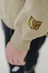 Canguro beige con capucha, cierre frontal parcial y bolsillo tipo canguro. Presenta estampado con texto en el pecho y detalle en la manga.