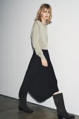 Jersey de punto color beige claro, con cuello en V y manga larga. Presenta detalles de botones decorativos en los hombros y en el ruedo inferior, sugiriendo un diseño desmontable o con aberturas ajustables.