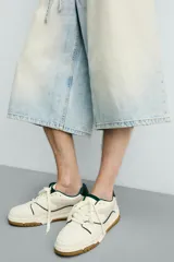Zapatilla deportiva de estilo retro con diseño de paneles en tonos crema y blanco, detalles en verde oscuro y suela de goma color caramelo.