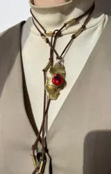 Collar artesanal con dije de base metálica irregular y piedra central roja. Presenta una estructura de tiento de gamuza marrón con detalles de tubos metálicos dorados.