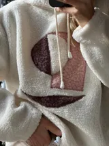 Buzo tipo hoodie de corderito color manteca, con capucha y cordones ajustables. Presenta un diseño abstracto frontal con apliques de textura suave en tonos rosa viejo y marrón.