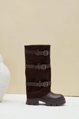 Bota alta de caña ancha, combinada en cuero ecológico y gamuza color marrón, con suela track gruesa y tres tiras horizontales con hebillas metálicas.
