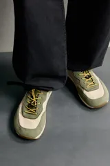 Zapatilla deportiva barefoot fabricada en combinación de piel y tejido, en tonos verde oliva y beige, con cordones elásticos amarillos y suela de goma flexible.