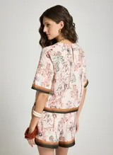 Blusa estampada Le Lis Blanc con diseño de paisaje tropical y arquitectónico, cuello redondo y mangas cortas con ribetes en tonos marrón y negro.