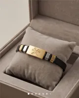 Pulsera de diseño profesional con placa central rectangular de acero dorado que presenta un grabado de tijeras. La estructura combina la placa con segmentos de cuerina negra y detalles metálicos, ofreciendo un estilo moderno y temático.