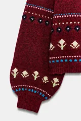 Jersey de punto con cuello subido y manga larga con puños ajustados. Presenta un diseño jacquard con motivos de lazos en color azul y detalles florales en tonos crema sobre una base de color bordó.