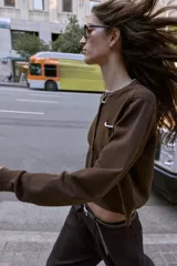 Chaqueta de punto color marrón con cuello redondo, manga larga y cierre frontal con botones. Presenta un bolsillo delantero tipo plastrón y detalles de ribetes en color celeste en el cuello y el bolsillo.
