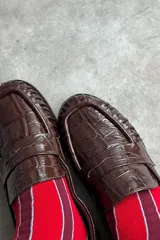 Mocasín de cuero marrón con textura croco y acabado brillante, de diseño clásico con detalle de antifaz en el empeine y costuras pespunteadas alrededor de la puntera.