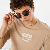 Remera color beige con estampado blanco en el frente que incluye un diseño gráfico de sol, olas y palmeras, y el texto 'Chill Zone Ahead'.