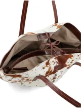 Bolso tipo tote de piel de vaca, con estampado moteado en blanco y marrón. Presenta asas de cuero marrón oscuro y costuras decorativas en forma de X en el centro frontal.