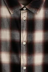 Camisa de manga larga confeccionada en franela de algodón suave, con diseño de cuadros en tonos marrones y beige. Presenta cuello inglés, cierre frontal con botones y bajo redondeado.