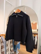 Sweater de punto color negro, con cuello polo y cierre frontal parcial, de manga larga y diseño holgado con terminaciones acanaladas en puños y cintura.