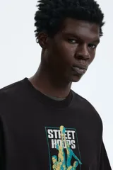 Remera de algodón marrón oscuro con cuello redondo y mangas cortas. Presenta un estampado frontal con gráficos de básquetbol y texto 'Street Hoops 95'.