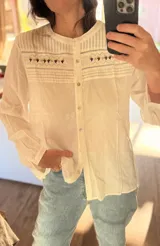 Blusa de manga larga color blanco con cuello redondo y cierre frontal de botones. Presenta detalles de bordado estilo punto smock en el pecho y textura de rayas verticales en la parte superior. Diseño holgado y estilo bohemio.