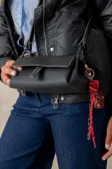 Cartera tipo bandolera de cuero sintético negro, con diseño rectangular, solapa frontal con cierre de broche y compartimento principal con cierre metálico. Incluye una correa larga ajustable y un detalle de llavero colgante con borlas y dijes.