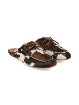 Zuecos Arno destalonados confeccionados en cuero con pelo con estampado animal print tipo vaca en tonos marrones, negros y blancos. Presentan diseño de inspiración náutica con cordones de cuero cruzados y ojalillos metálicos dorados.