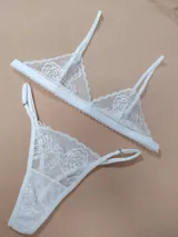 Conjunto de lencería de dos piezas confeccionado en encaje blanco con diseño floral. Incluye un sostén triangular con tirantes regulables y una bombacha colaless ajustable con tiras laterales.