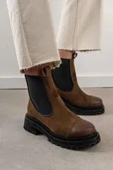 Bota tipo Chelsea de cuero marrón con suela track negra de goma. Presenta paneles elásticos laterales y una costura reforzada en la puntera. Incluye una correa de cuero desmontable con hebilla metálica.