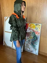 Piloto impermeable de color verde oliva con detalles de volados en color naranja sobre los hombros. Presenta capucha integrada, cintura ajustable mediante cordón con tranca, mangas voluminosas con puños elastizados y bolsillos frontales con solapa.