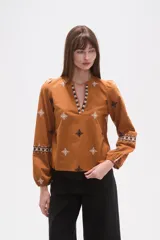 Blusa de algodón color tostado con cuello mao y escote en V. Presenta mangas largas abullonadas con puños elásticos y un diseño con bordados geométricos en contraste distribuidos por toda la prenda.