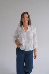 Blusa blanca de manga larga con diseño de rayas verticales y bordados florales en tono grisáceo. Presenta cuello camisero, cierre frontal con botones y puños elásticos.