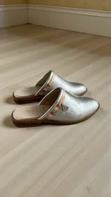 Sandalias chatitas tipo mules, color plateado con detalle de arte en la parte superior y aplique metálico triangular.