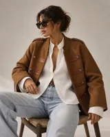 Chaqueta de estilo militar color marrón, confeccionada en tejido de lana con textura. Presenta detalles en cuero en los hombros y en las solapas frontales, acompañados de botones metálicos decorativos.