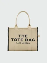 Bolso tipo tote de gran tamaño en color beige, confeccionado en lona resistente. Presenta el logo estampado en negro en el frente y detalles de cintas laterales con inscripciones. Cuenta con doble asa superior para llevar al hombro.