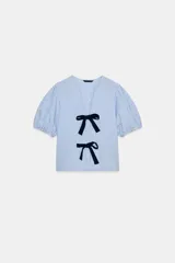 Blusa de cuello redondo y escote pico con manga corta abullonada acabada en elástico. Detalle de lazos a contraste en delantero. Bajo recto.