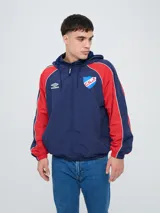 Campera deportiva con capucha Umbro del Club Nacional de Football, modelo Venti. Diseño en color azul marino con mangas rojas y detalles de ribetes blancos, cierre frontal completo y bolsillos laterales. Cuenta con el escudo del club y el logo de la marca bordados en el pecho.