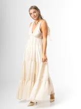 Vestido largo color crudo, confeccionado en mezcla de tencel y lino, con escote en V pronunciado, breteles anchos terminados en lazos en los hombros, y falda larga de varios niveles con volados amplios y fluidos.